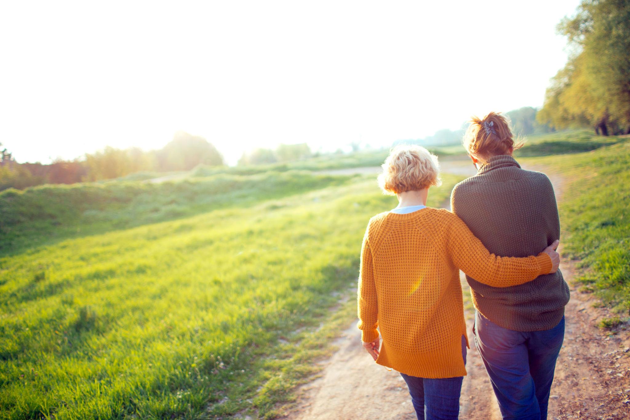 Dos mujeres, una más alta que la otra caminando por un camino de tierra con grama o pasto a ambos lados del camino de espaldas a la cámara.